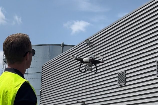 Un homme en gilet jaune observe un drone volant près d'un bâtiment industriel lors d'une opération de dépigeonnage par effarouchement.